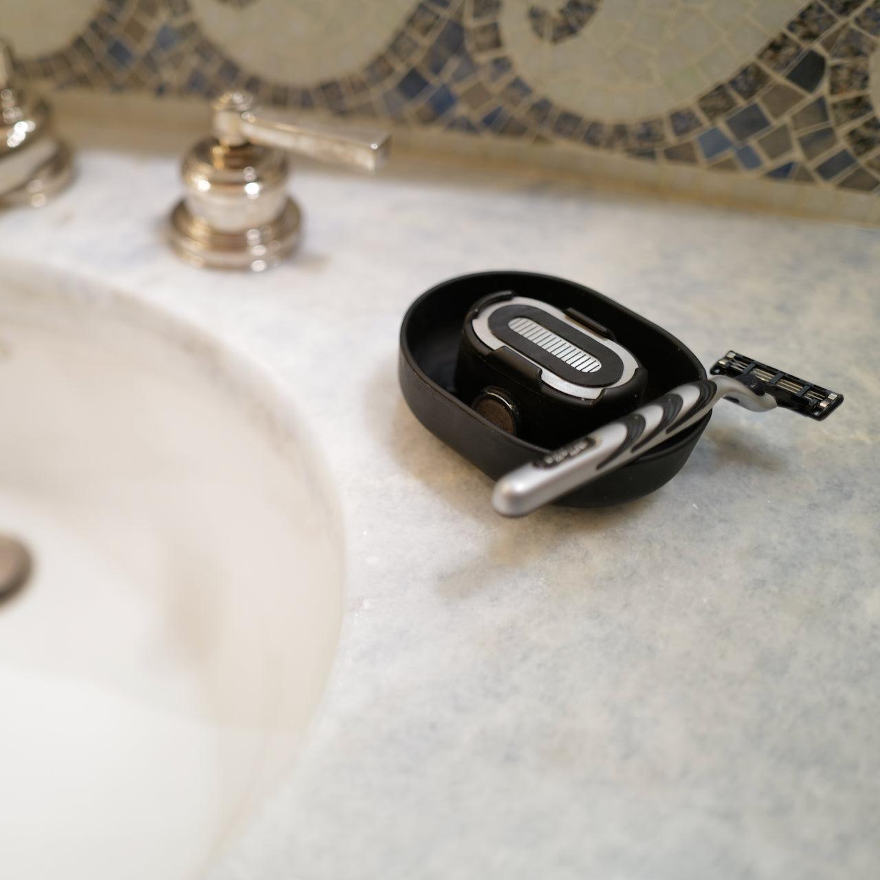 A black Razor Rinser, which shoots water jets to unclog your razor, situated on a bathroom countertop next to a sink.