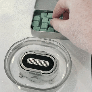 A hand drops a shave bomb from the tin into the Razor Rinser®, where it fizzes into foam